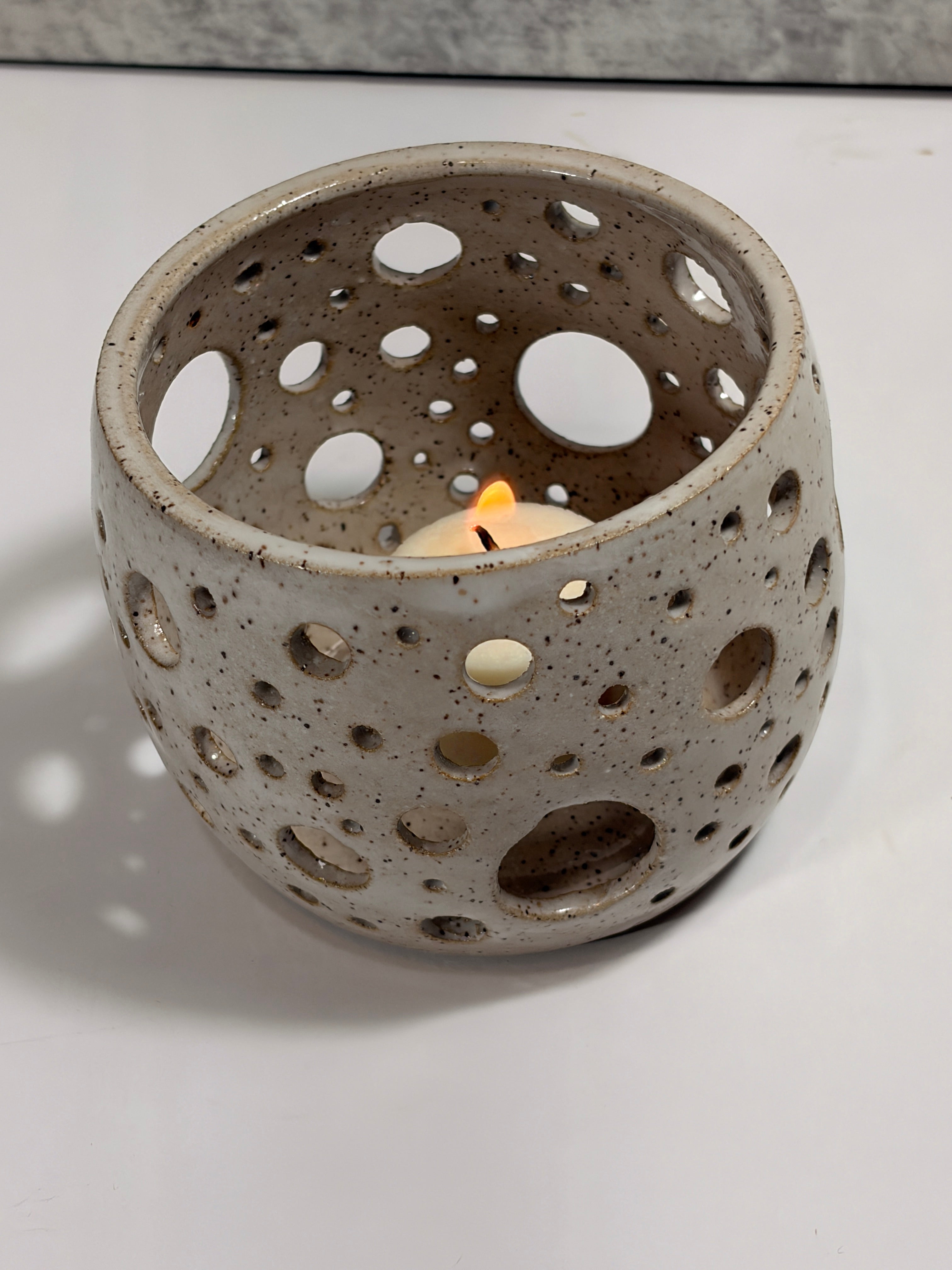 A speckled ceramic candle holder with a lit tea light candle inside, set on a white surface with a soft shadow.
