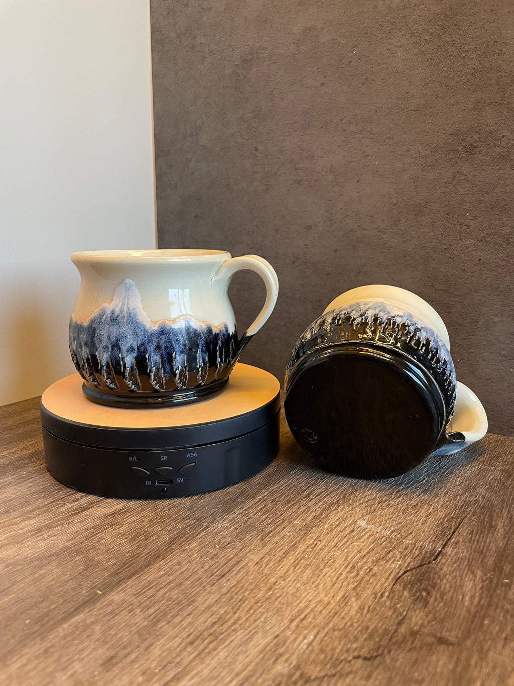 A ceramic mug with a mountain landscape design in blue, black, and cream, shown on a wooden surface with its black lid beside it.