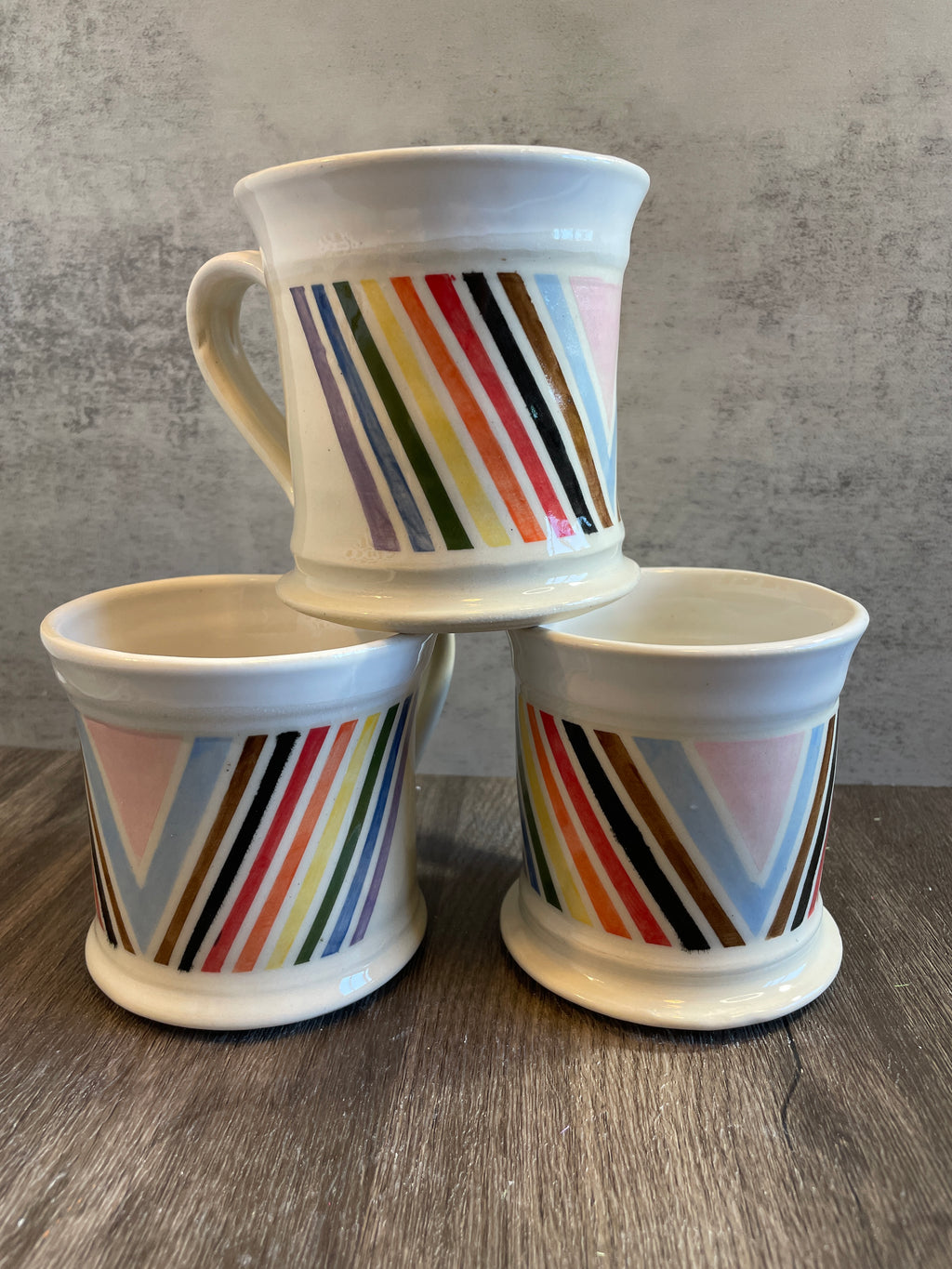 Three white ceramic tumblers with colorful diagonal striped patterns, arranged in a pyramid stack on a wooden surface against a gray textured wall.