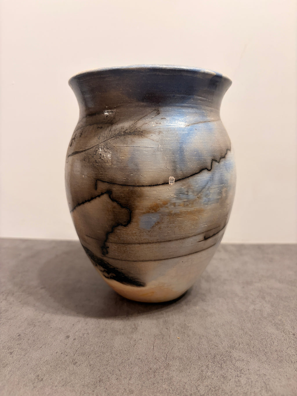 A handmade ceramic vase with a rounded body, flared rim, and textured glaze featuring blue, gray, brown, and black marbled patterns, set against a neutral beige and gray background.