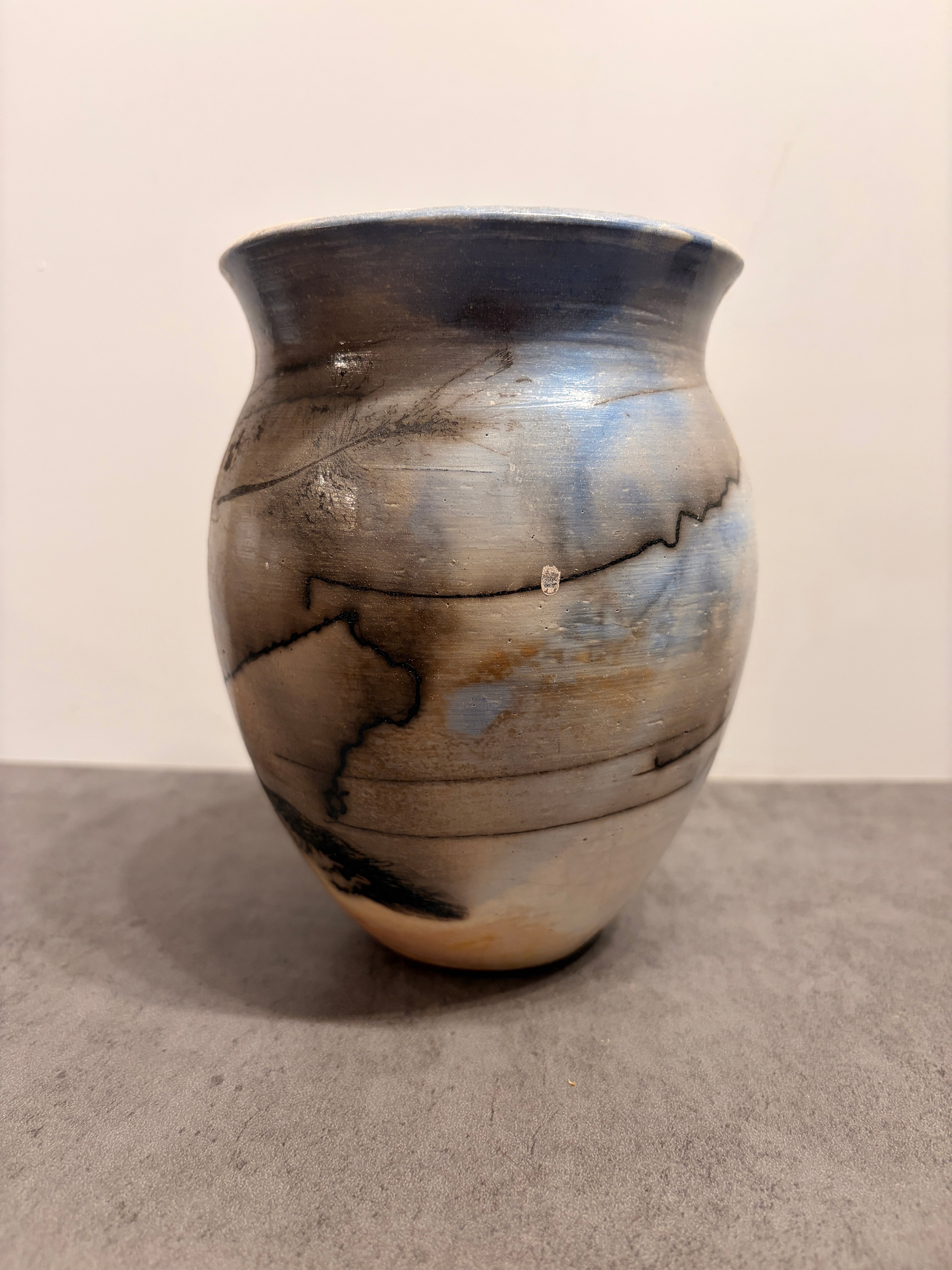 A handmade ceramic vase with a rounded body, flared rim, and textured glaze featuring blue, gray, brown, and black marbled patterns, set against a neutral beige and gray background.