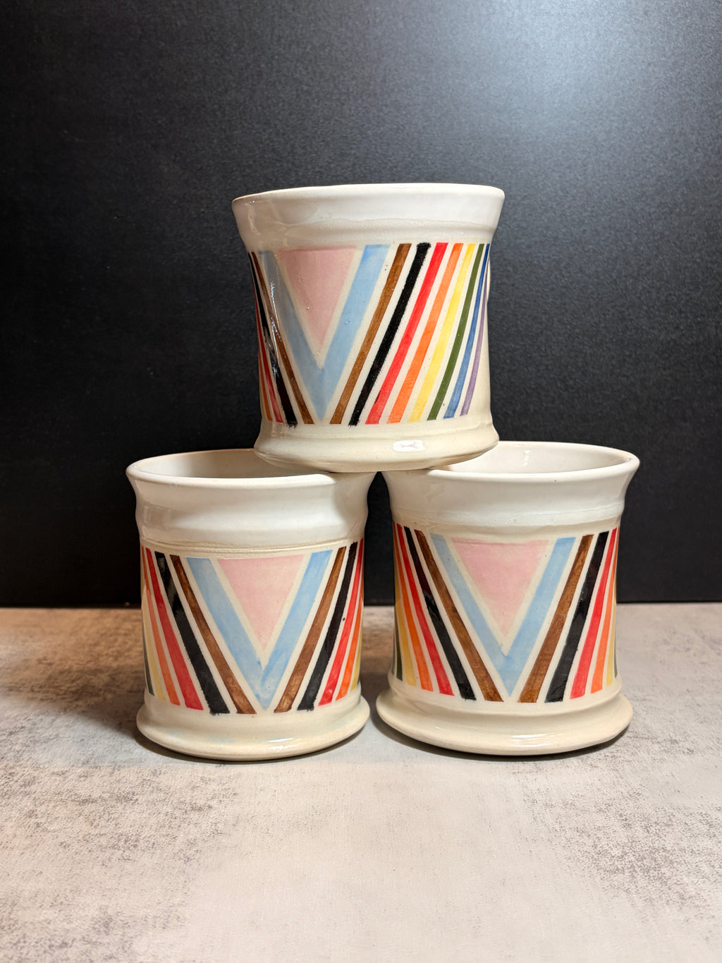 Three ceramic toothbrush holders stacked in a pyramid arrangement, featuring a colorful retro chevron stripe pattern on a cream-colored glaze, set against a dark gray background.