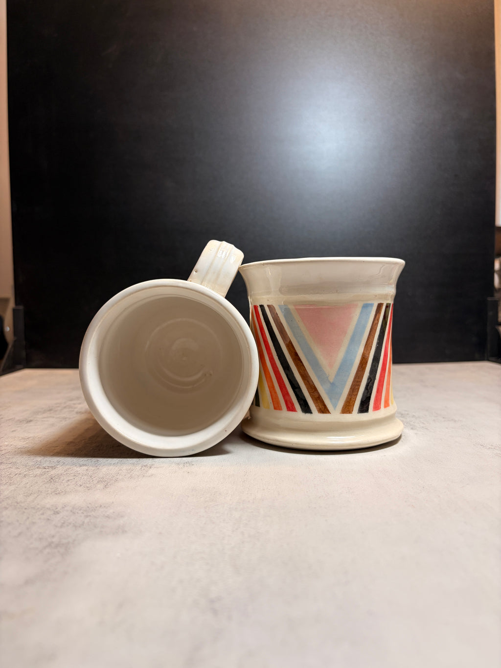 A ceramic mug with a matching lid, displayed on a light gray surface against a dark background. The mug features a cream-colored body with a colorful geometric chevron pattern on the side.