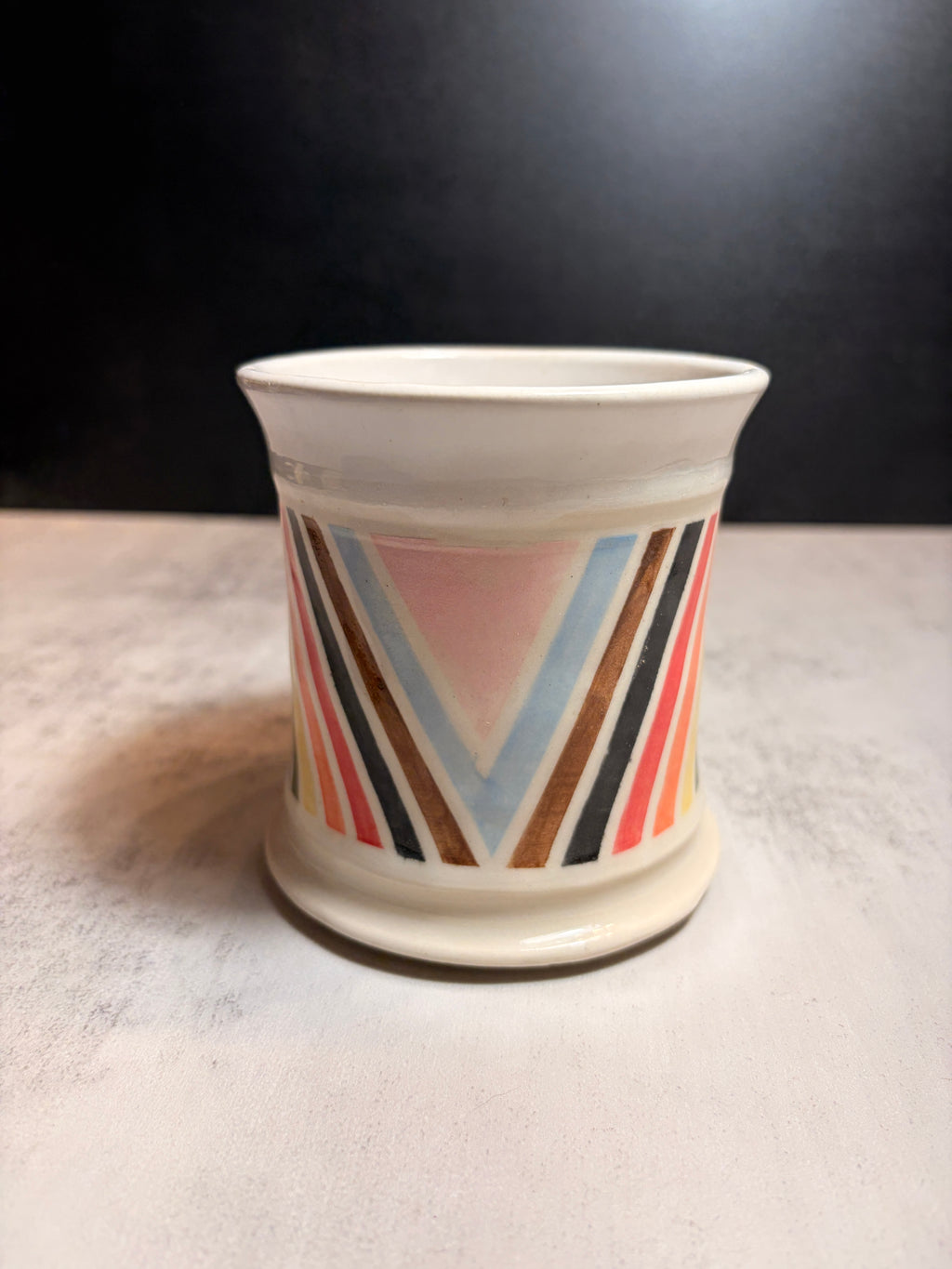 A small ceramic cup with a white base and a colorful geometric pattern featuring pink, blue, orange, brown, and black stripes, set on a light gray surface against a dark background.