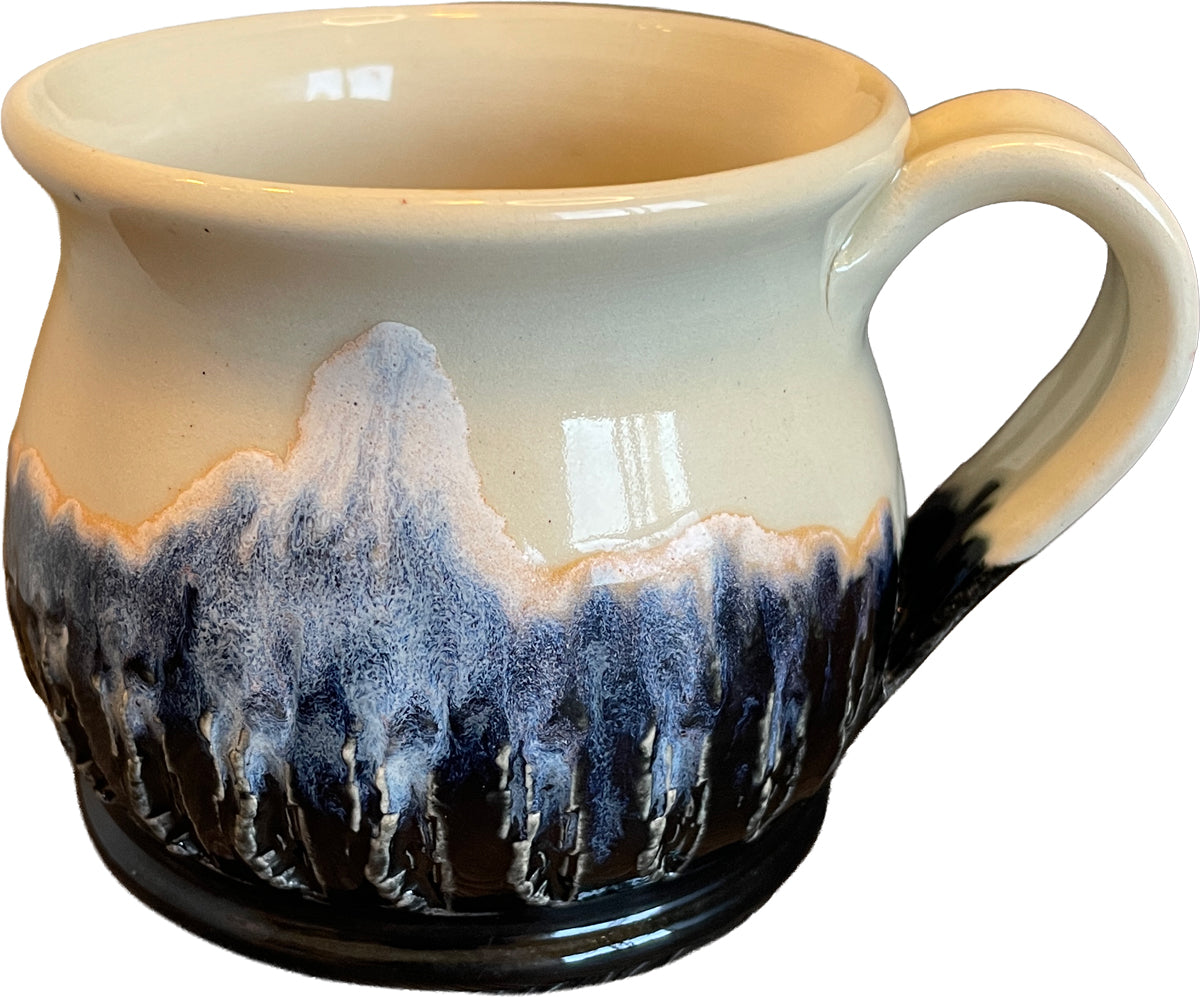 A ceramic mug with a cream-colored glaze and a dark blue to black dripping mountain-like pattern around the lower half, shown on a plain white background.