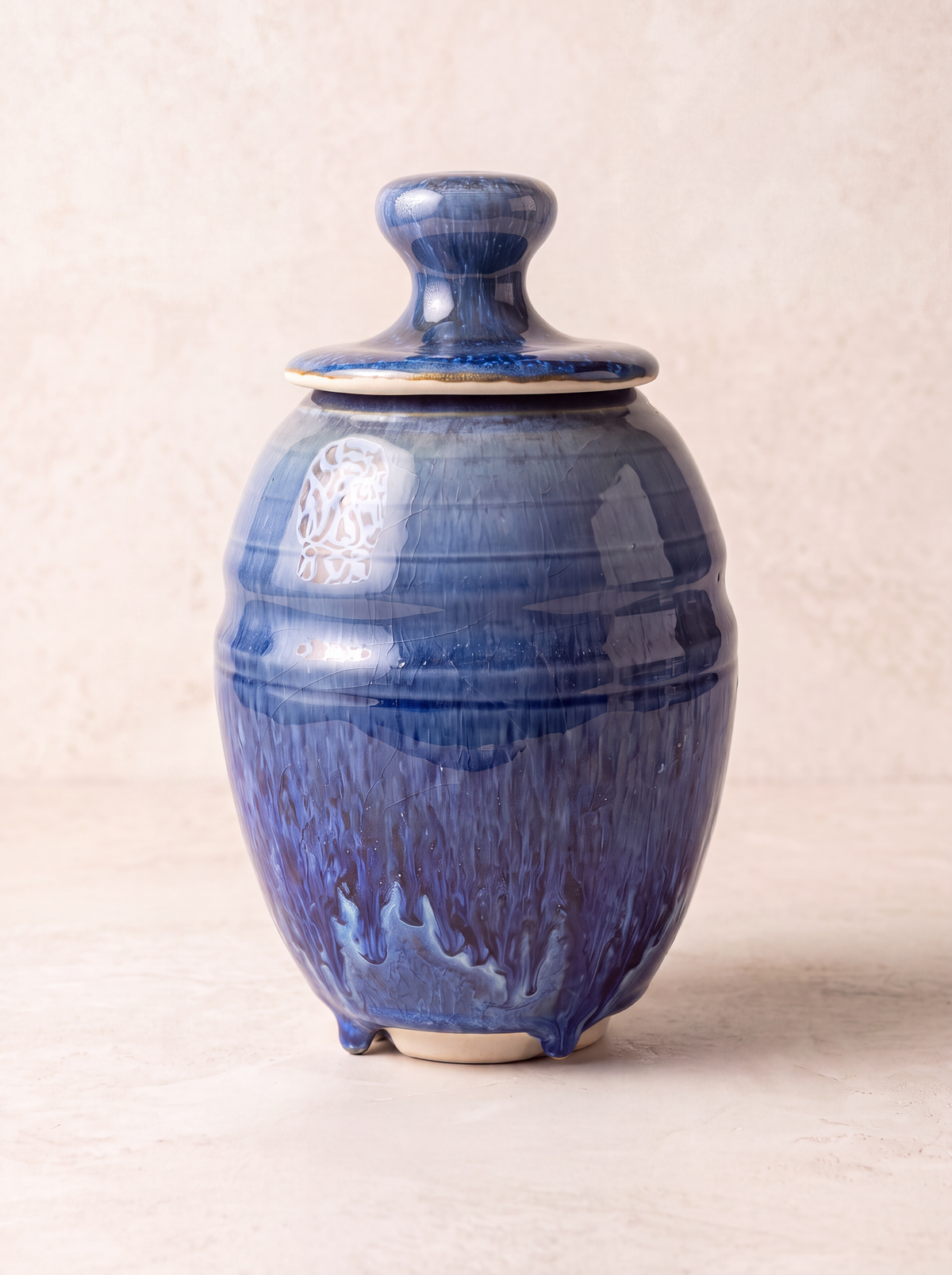 A glazed ceramic lidded jar in deep cobalt blue with a textured, dripping finish and a small white decorative mark on the side, set against a neutral beige background.
