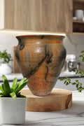A handmade ceramic planter with a textured, earthy glaze in shades of brown, orange, and black, displayed on a round wooden riser in a modern kitchen setting.