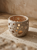 A small speckled ceramic candle holder with circular cutouts, lit with a warm candlelight glow, set on a textured stone surface against a rustic beige background.