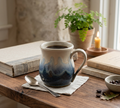 A ceramic mug filled with coffee sits on a wooden table in a cozy indoor setting, surrounded by books, a spoon, coffee beans, and a potted plant in the background.